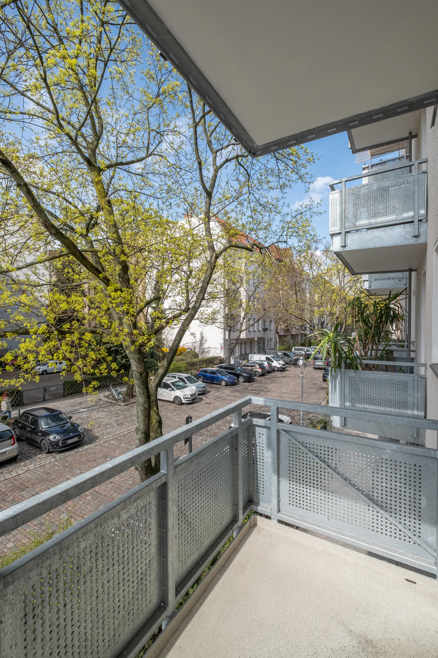 Balcony view with metal railings overlooking a parking lot with cars and a tree with green leaves.