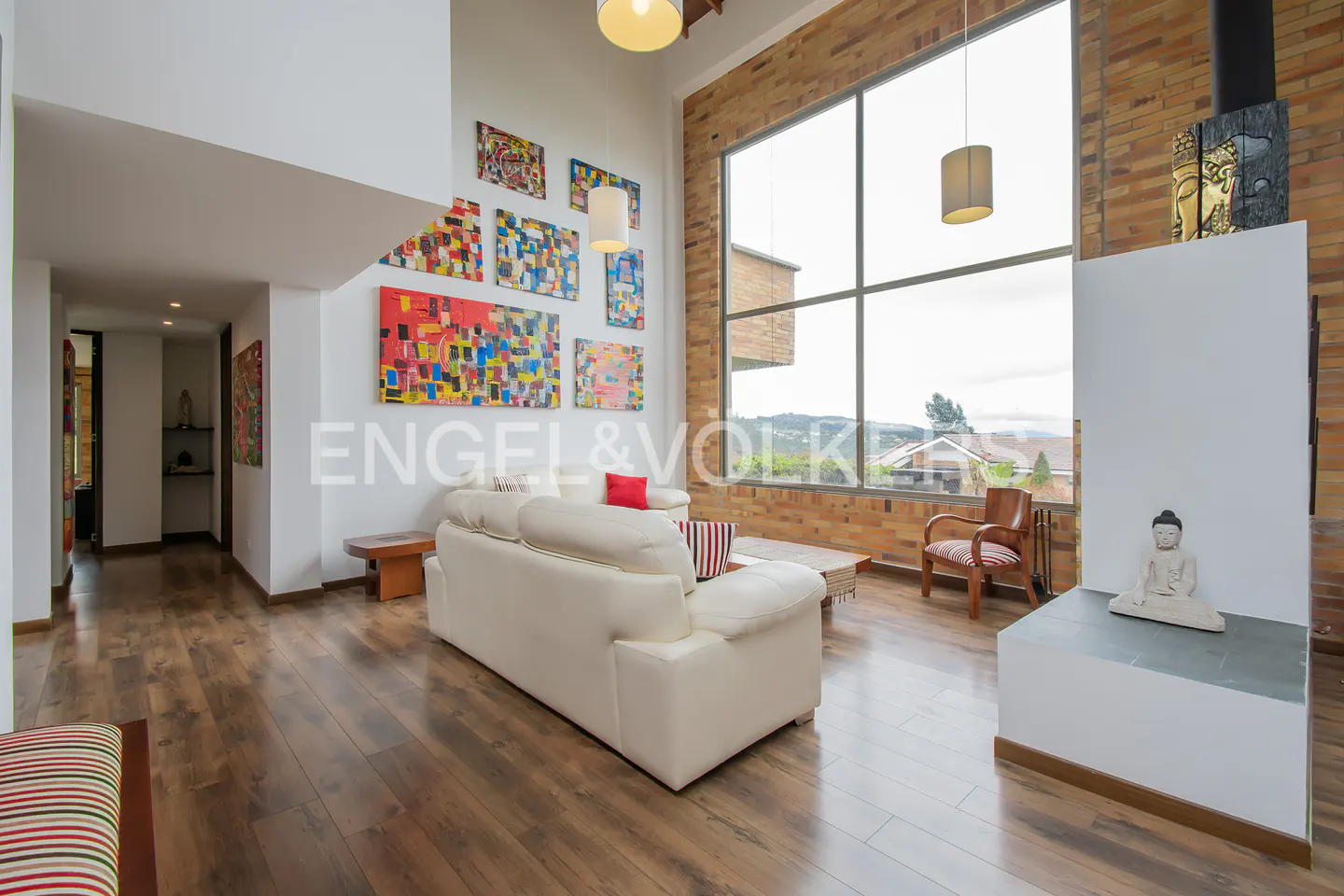 Bright living room with hardwood floors, white sofa, and colorful artwork. Large windows offer a view of the landscape. Buddha statue on a white fireplace.
