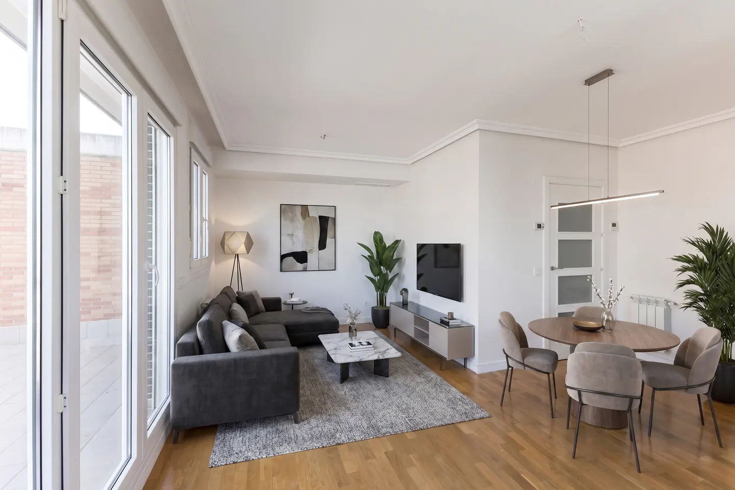 Bright living room with a gray sectional sofa, marble coffee table, and round dining table with four chairs. Large windows and wood floors.
