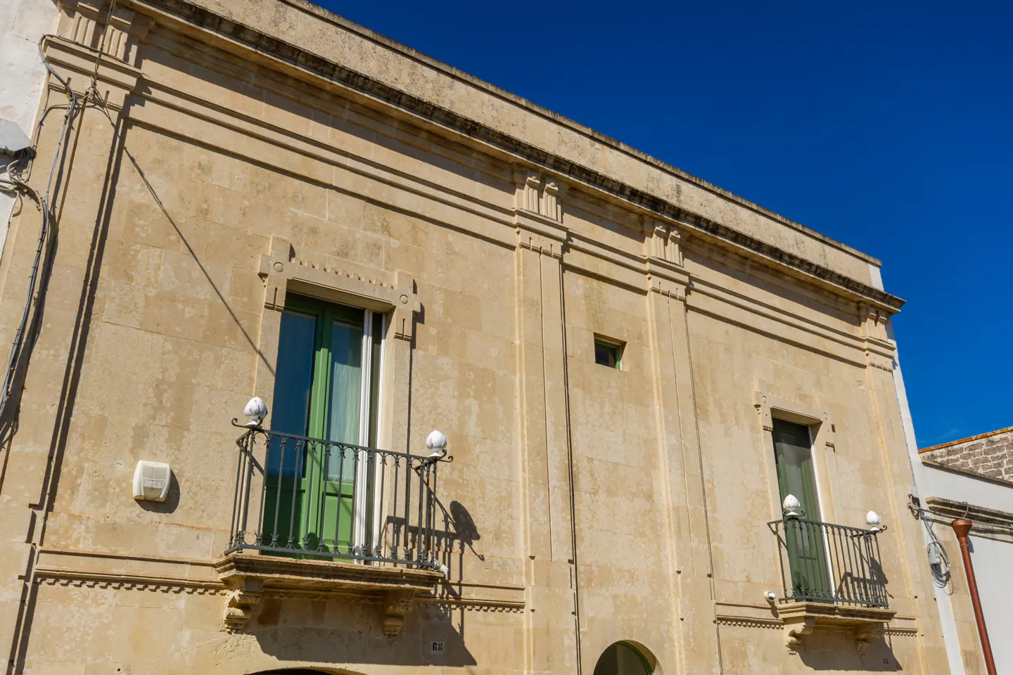 Two-story beige building with green-framed windows and black wrought-iron balconies against a clear blue sky.