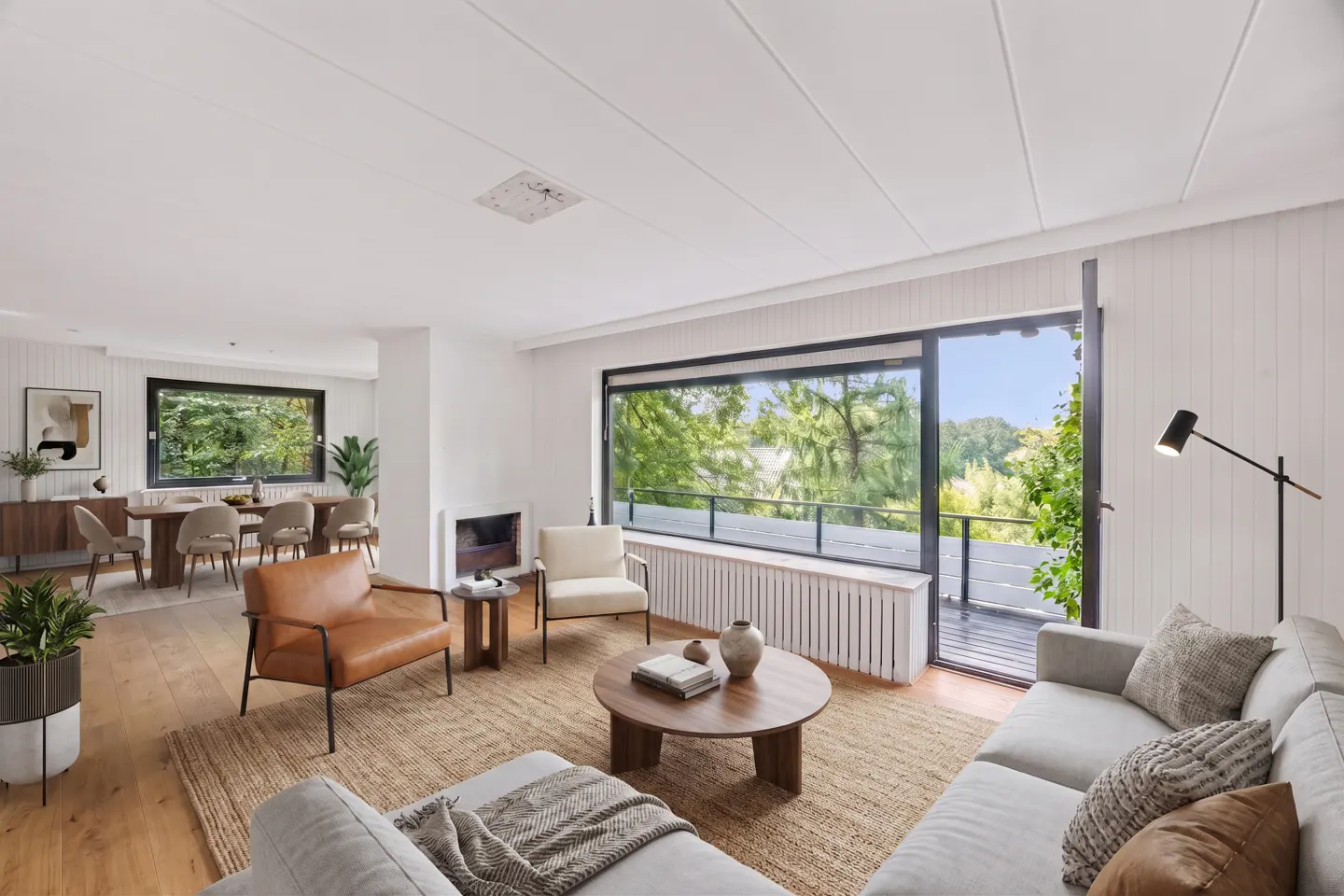 Bright, modern living room with a gray sofa, brown leather chair, and a round wooden coffee table on a jute rug. Large windows overlook green trees.