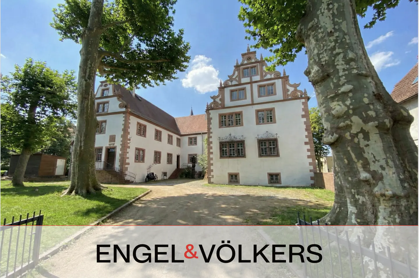 A historic white manor with ornate gables and brown trim, framed by large trees on a sunny day with a dirt driveway.