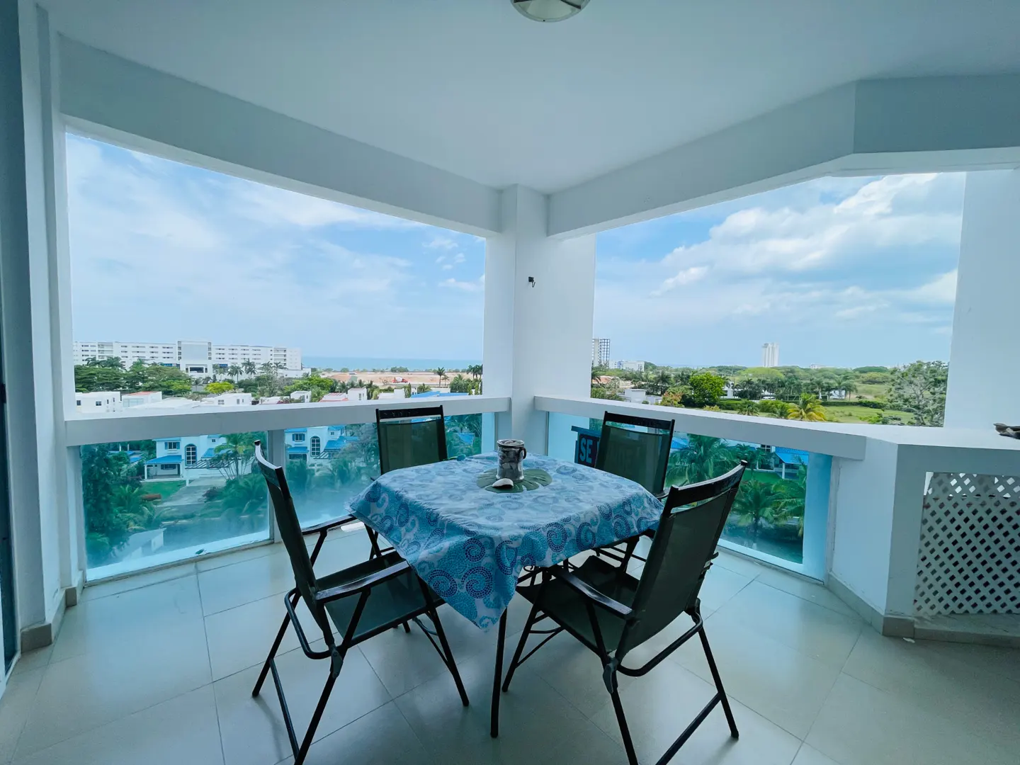 Balcony view with table and chairs. Blue patterned tablecloth, ocean view, and white building exterior.