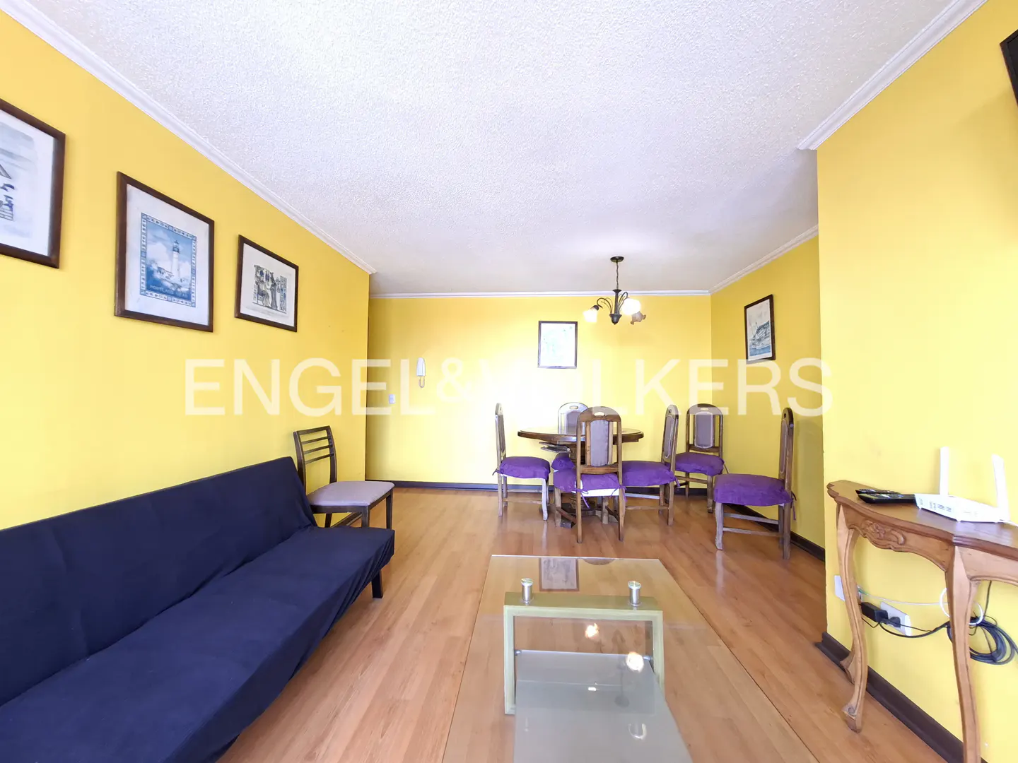 Bright living room with yellow walls, wood floors, and a blue sofa. A dining table with purple chairs sits in the center. Artwork adorns the walls.