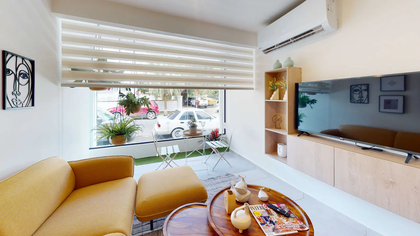 Bright living room with yellow sofa, wooden table, and large window. Outside, a small patio set and street view are visible.