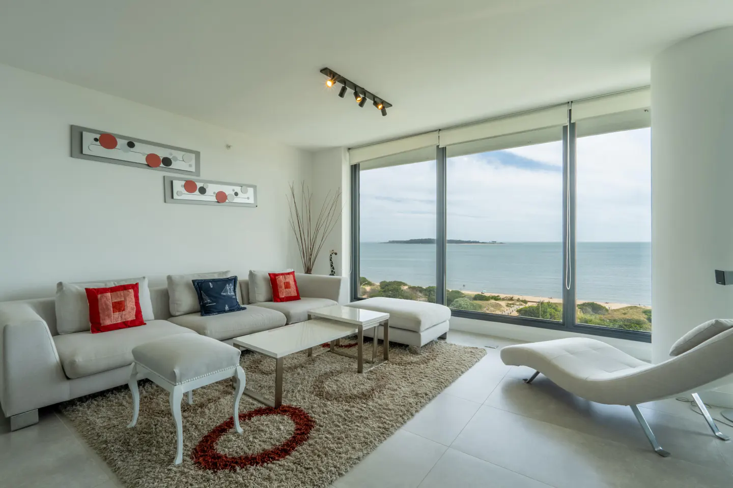 Modern living room with white sofa, chaise lounge, and coffee tables on a textured rug, facing a large ocean view window.