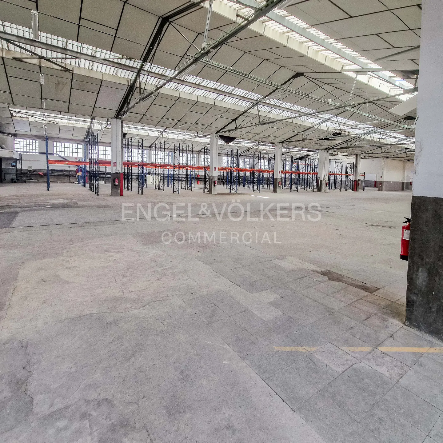 A large, empty warehouse with concrete floors and metal shelving units. The Engel & Völkers Commercial logo is visible.