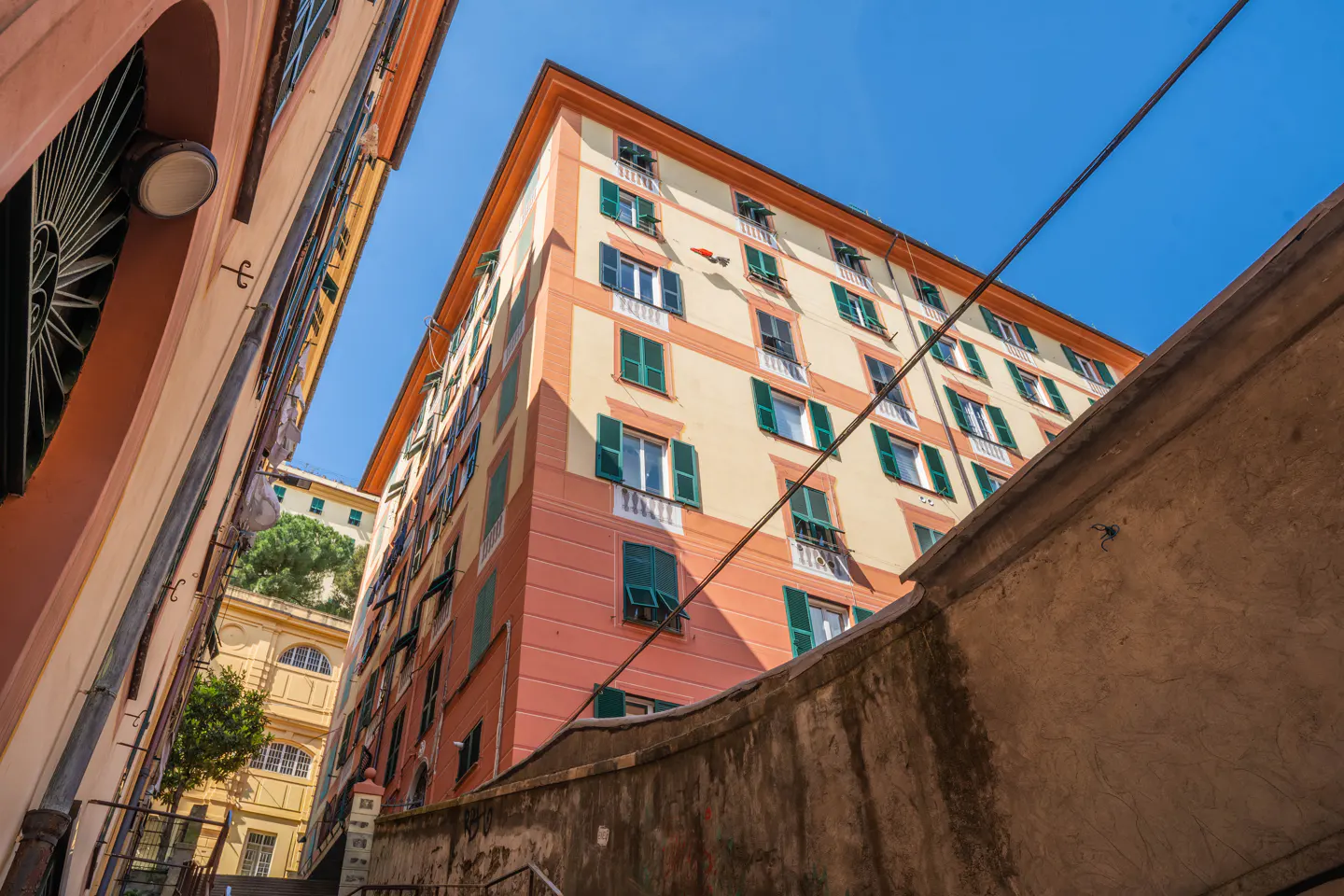 Looking up at a tall, peach-colored building with green shutters from a narrow street in Genoa, Italy.