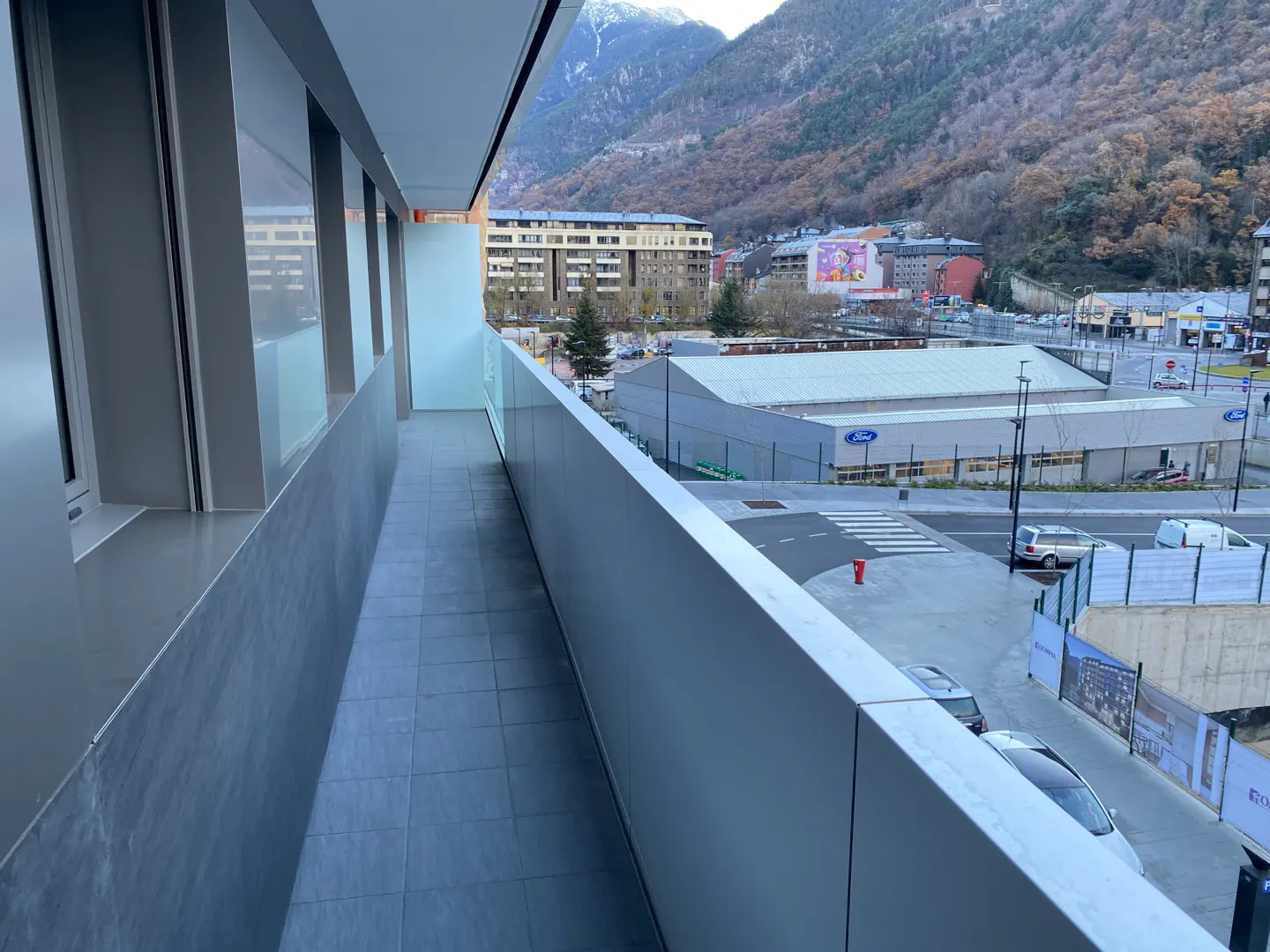Balcony view of a city with mountains in the background. Gray tiled floor and metal railing. Buildings and cars are visible below.