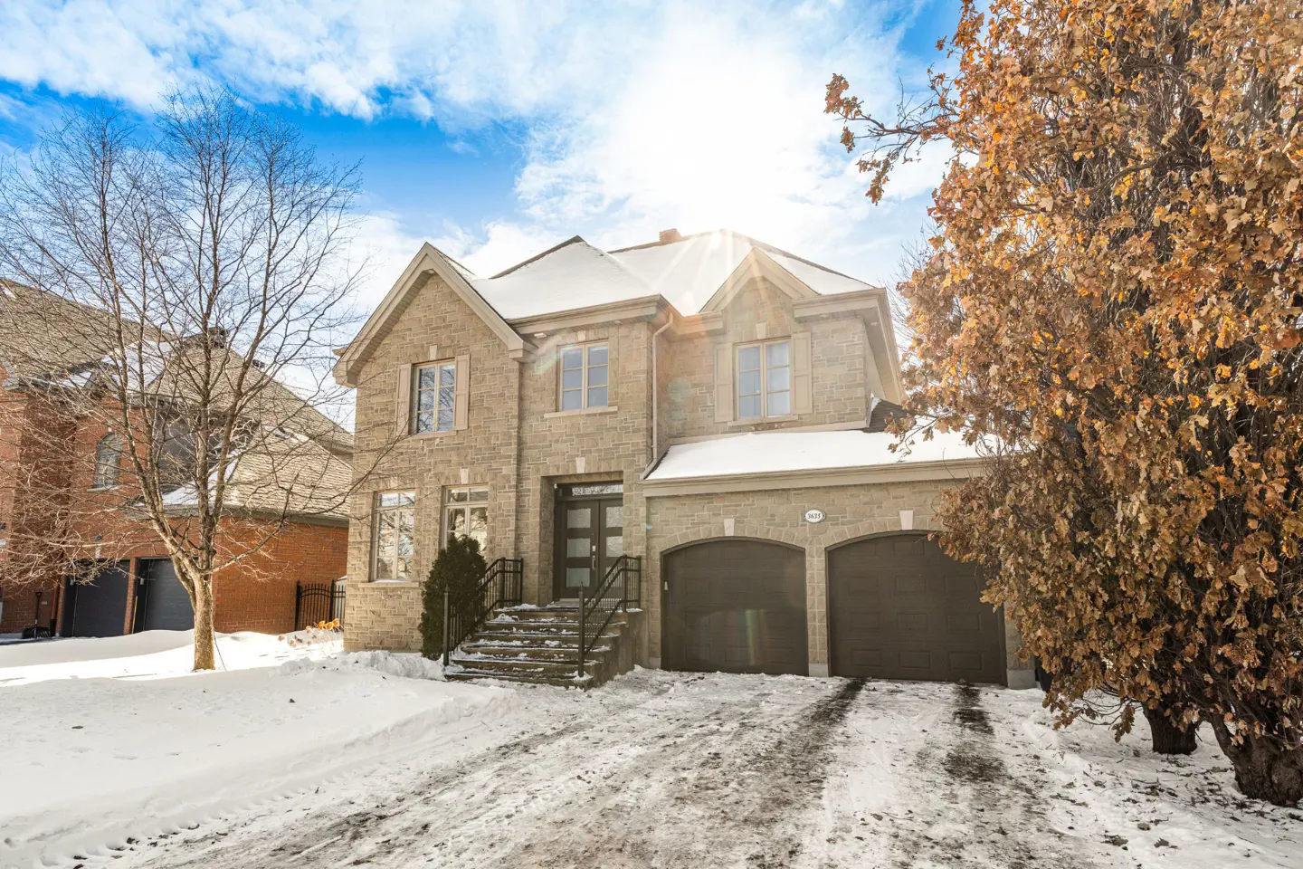 Two-story stone house with a two-car garage and snow-covered yard on a sunny day.