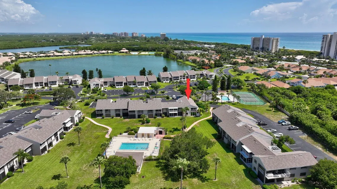 Aerial view of condos with a pool, tennis court, and lake. An arrow points to a specific unit. The ocean and city are in the background.