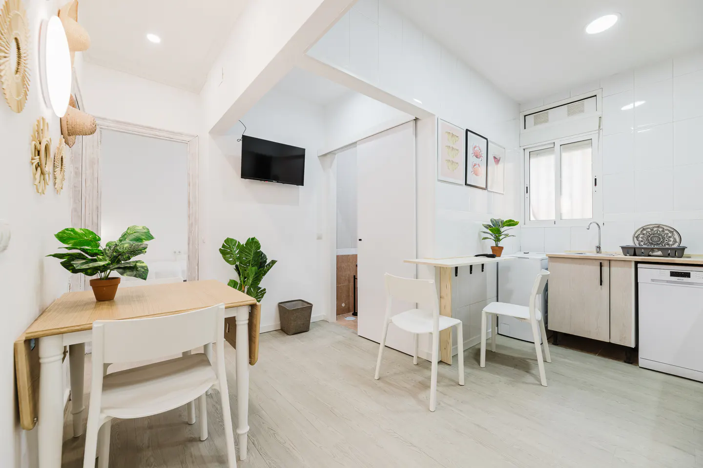 Bright, modern apartment interior with white walls, light wood floors, and a small kitchen area. Table with two chairs and a TV on the wall.