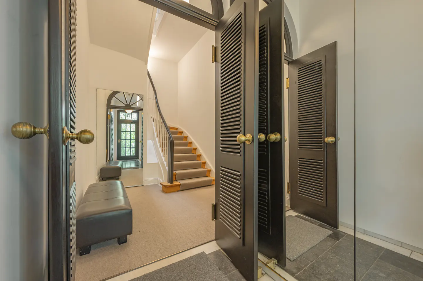 Open black louvered doors reveal a foyer with a carpeted staircase, bench seating, and a window with a view to the outside.