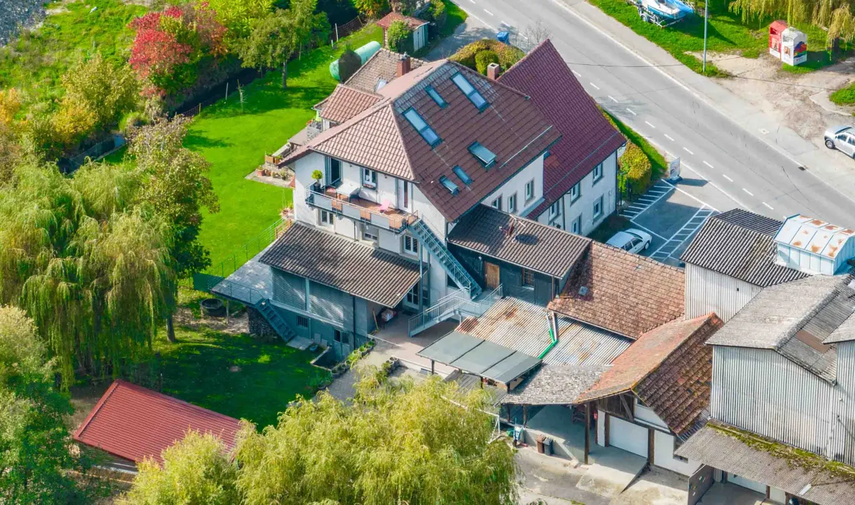 Aerial view of a multi-story white house with a red tile roof, surrounded by green trees and a lawn. A road runs alongside the property.