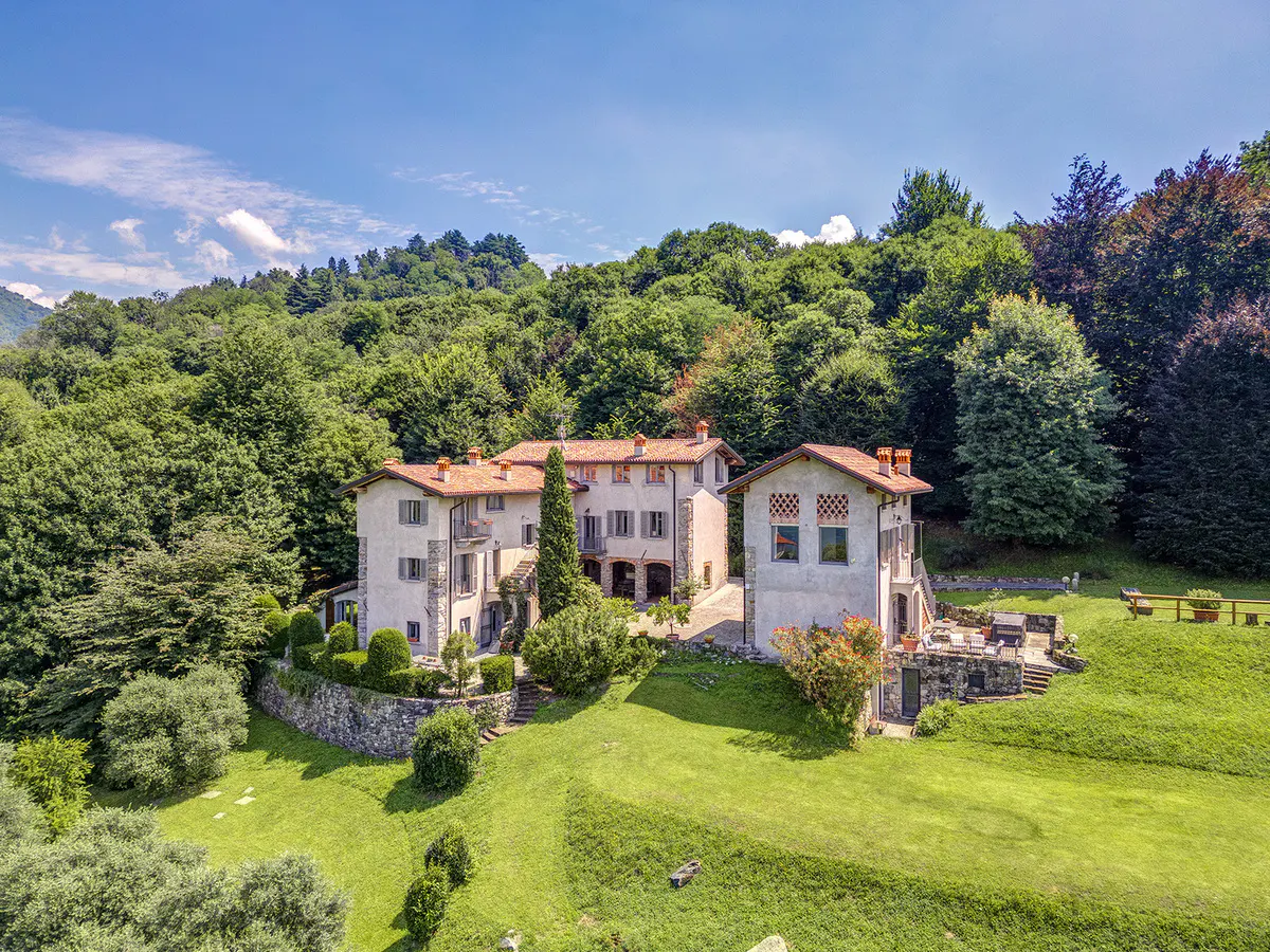 Aerial view of a large, light-stone house with a red tile roof, surrounded by green trees and a grassy lawn.