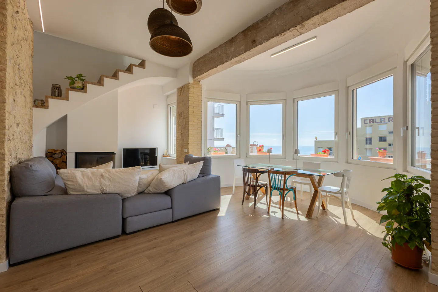 Bright living room with wood floors, a gray sofa, and a glass table with chairs. Large windows offer a sea view. Stairs lead up to another level.