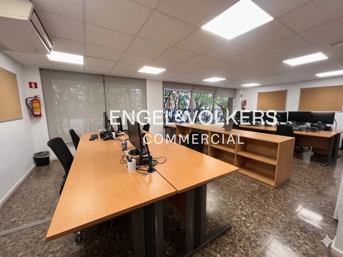 Bright office space with desks, computers, and chairs. Engel & Volkers Commercial logo overlay. Windows and shelving visible.