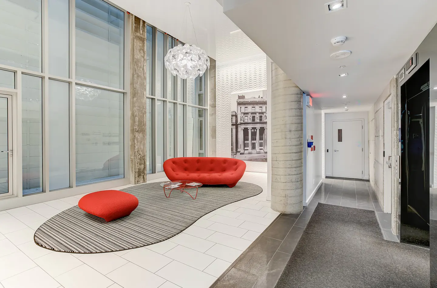 Bright lobby with large windows, red sofa and ottoman on a striped rug. Concrete pillars and a hallway with white doors are visible.