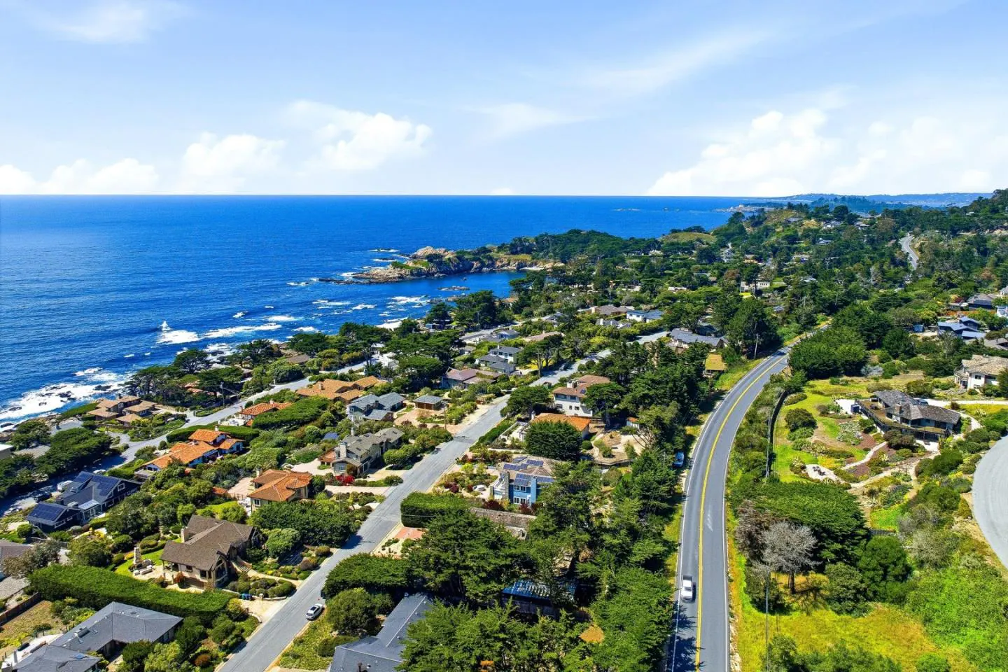 Aerial view of a coastal neighborhood with houses, trees, and a road next to the blue ocean under a bright sky.