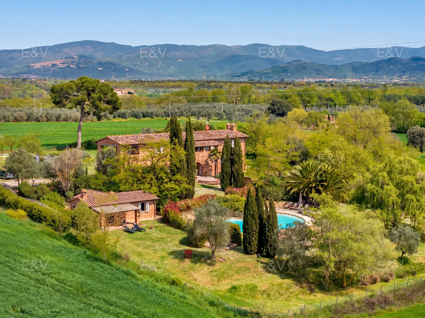 Tuscan villa with pool, surrounded by green fields and trees, under a blue sky. Mountains are visible in the background.