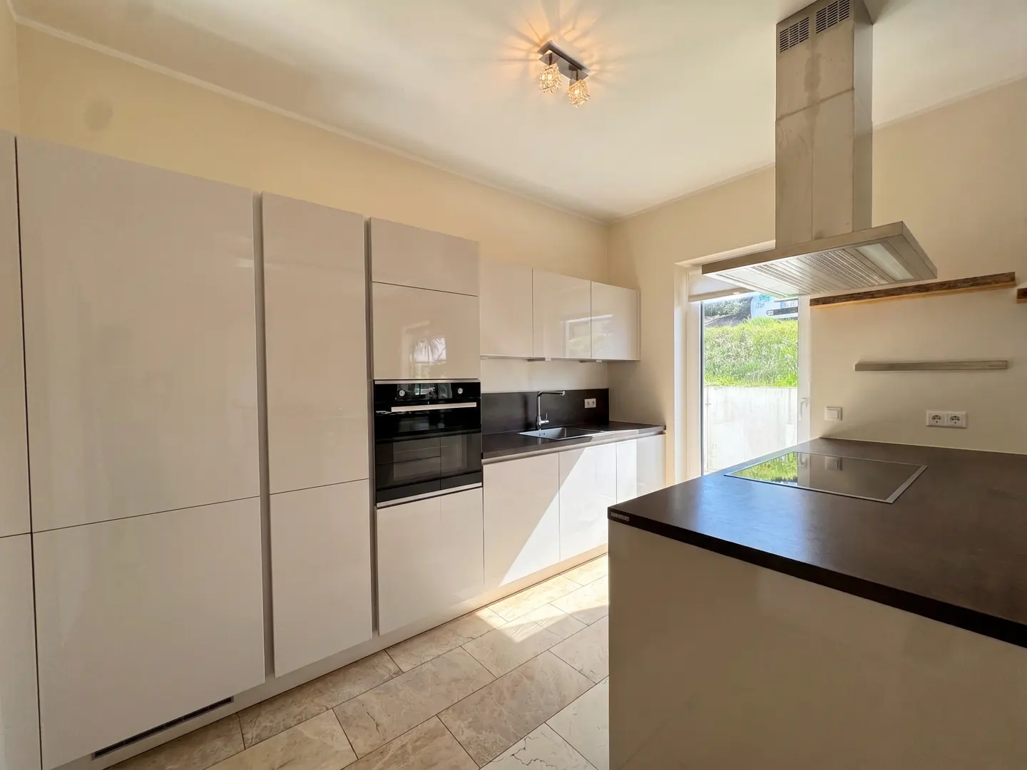 Bright kitchen with white cabinets, dark countertops, and a stainless steel range hood. A kitchen island with a cooktop is in the foreground.