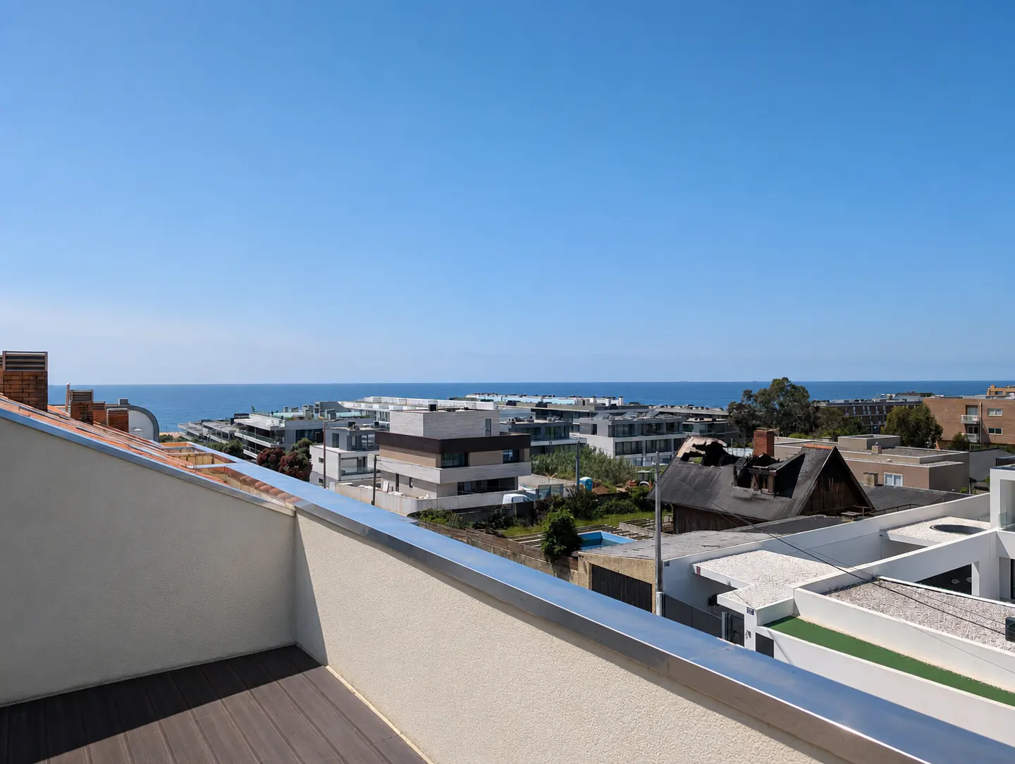 View from a balcony with wood floors, a white wall, and a metal railing overlooking houses and the ocean under a clear blue sky.