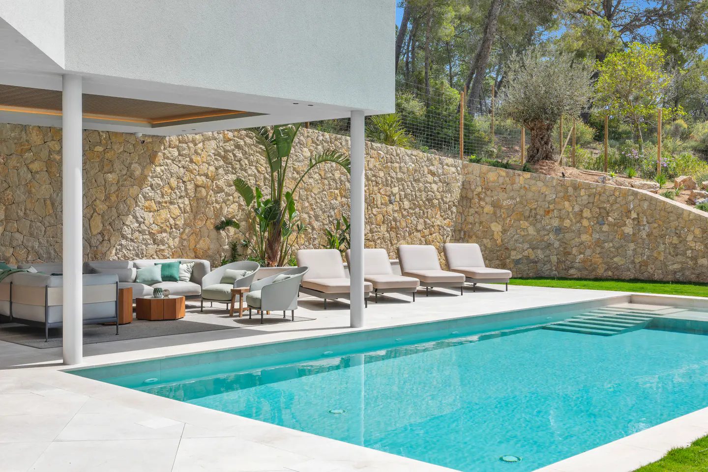 Outdoor patio with a pool, stone wall, and lounge furniture. The pool has steps leading into the water. Trees are visible in the background.