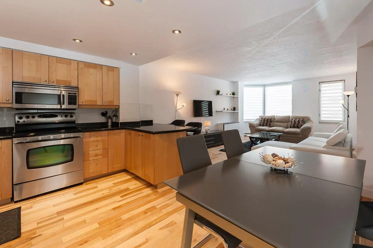 Open-concept living space with a kitchen featuring stainless steel appliances and light wood cabinets, a dining table, and a living room with sofas.