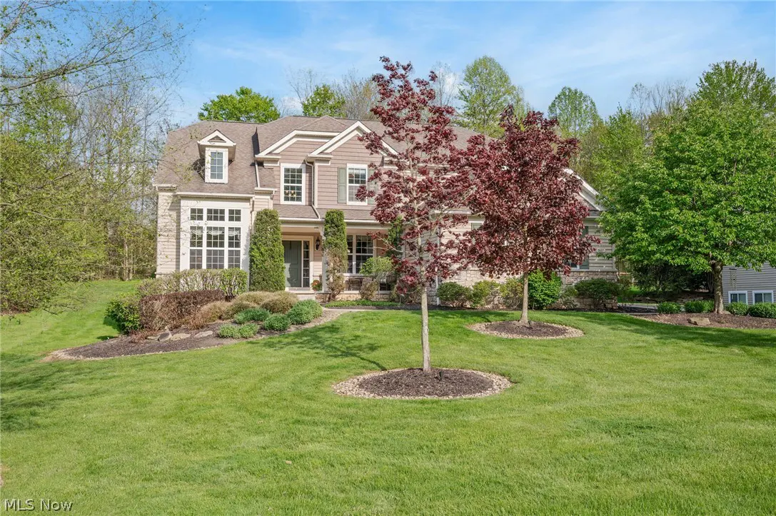 Two-story house with beige siding and stone accents, green lawn, and trees with red and green leaves.