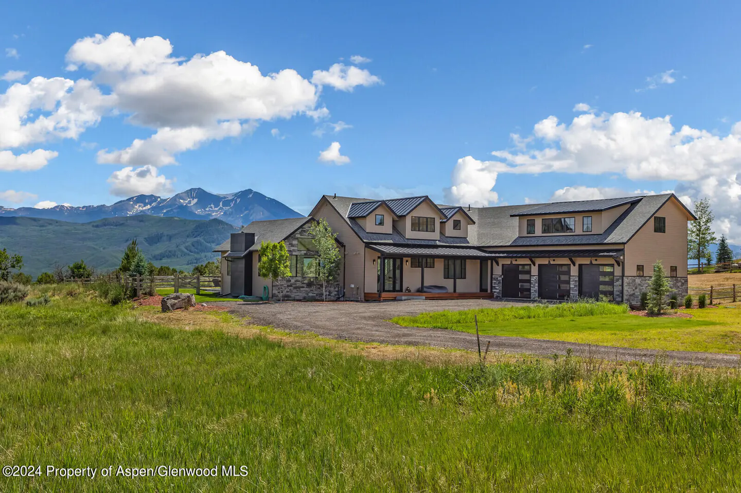 Exterior of a tan two-story house with a black roof and three-car garage. Mountains and a blue sky with clouds are in the background.