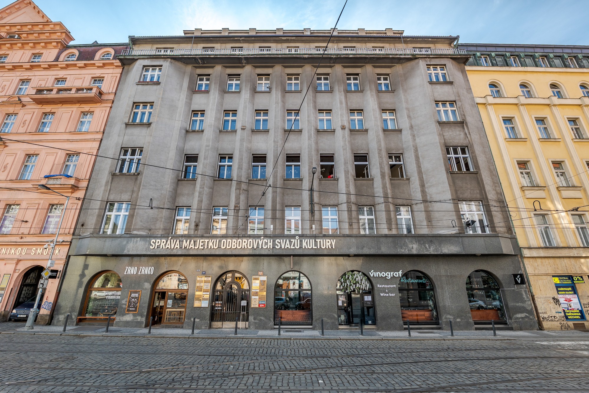 Street view of a gray building with arched storefronts and white windows, flanked by pink and yellow buildings.