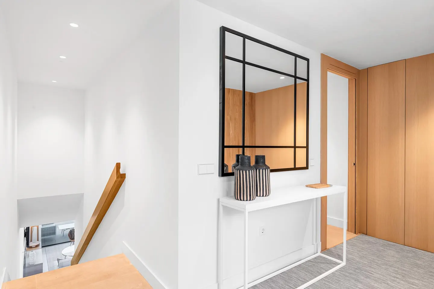 Bright hallway with white walls, wood stairs, and a black-framed mirror above a white console table with striped vases.