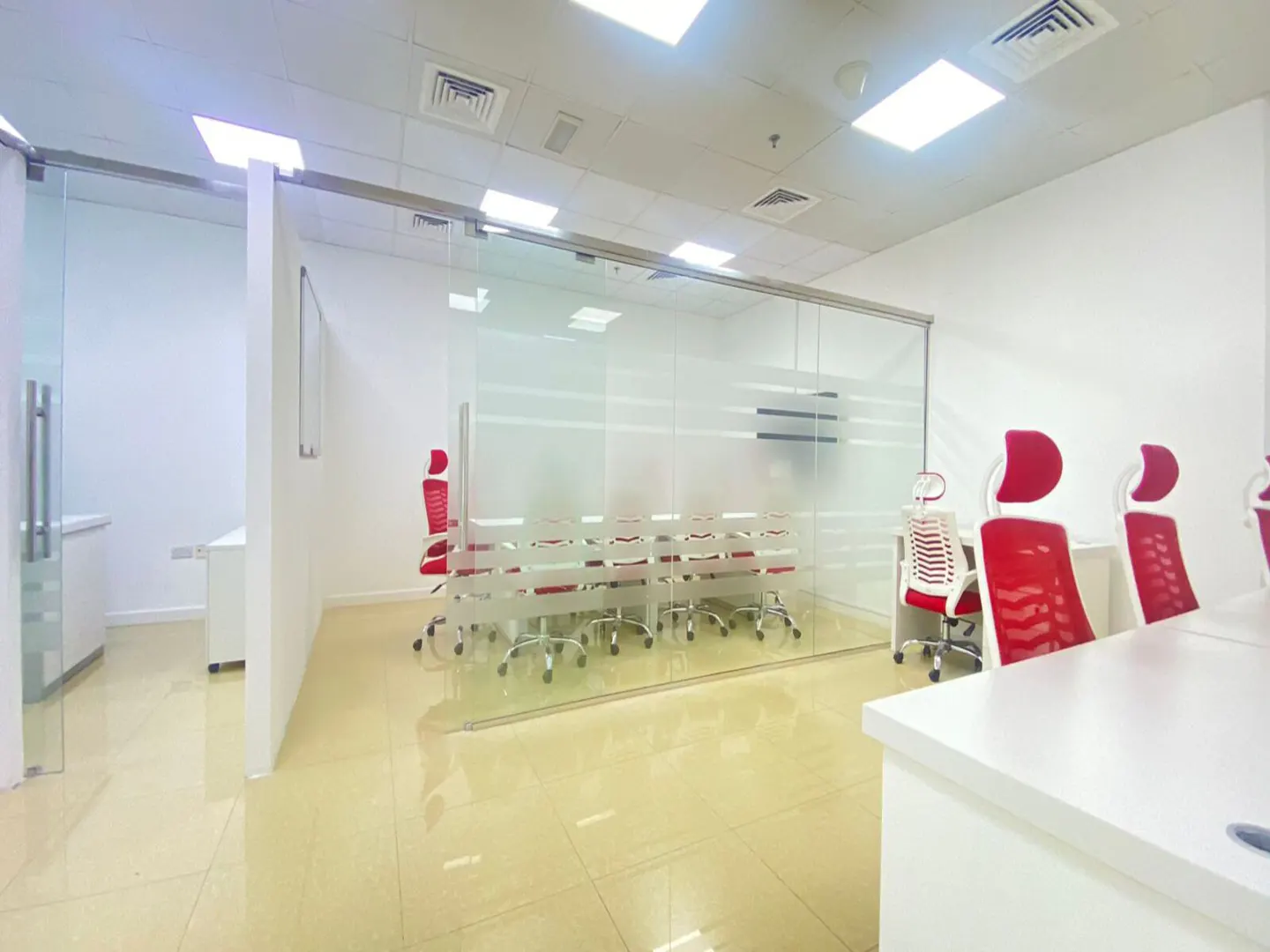 Bright office space with glass-walled conference room. Red chairs surround a long table. White desks and cabinets are visible.