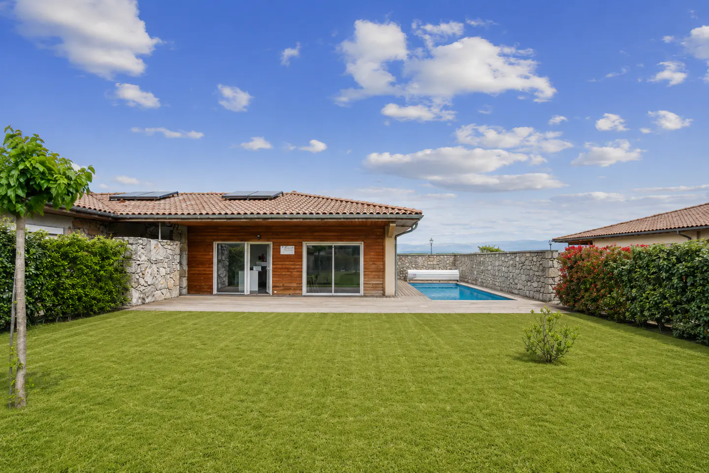 Backyard view of a wood-sided house with a red tile roof, green lawn, stone wall, and a blue swimming pool under a cloudy sky.