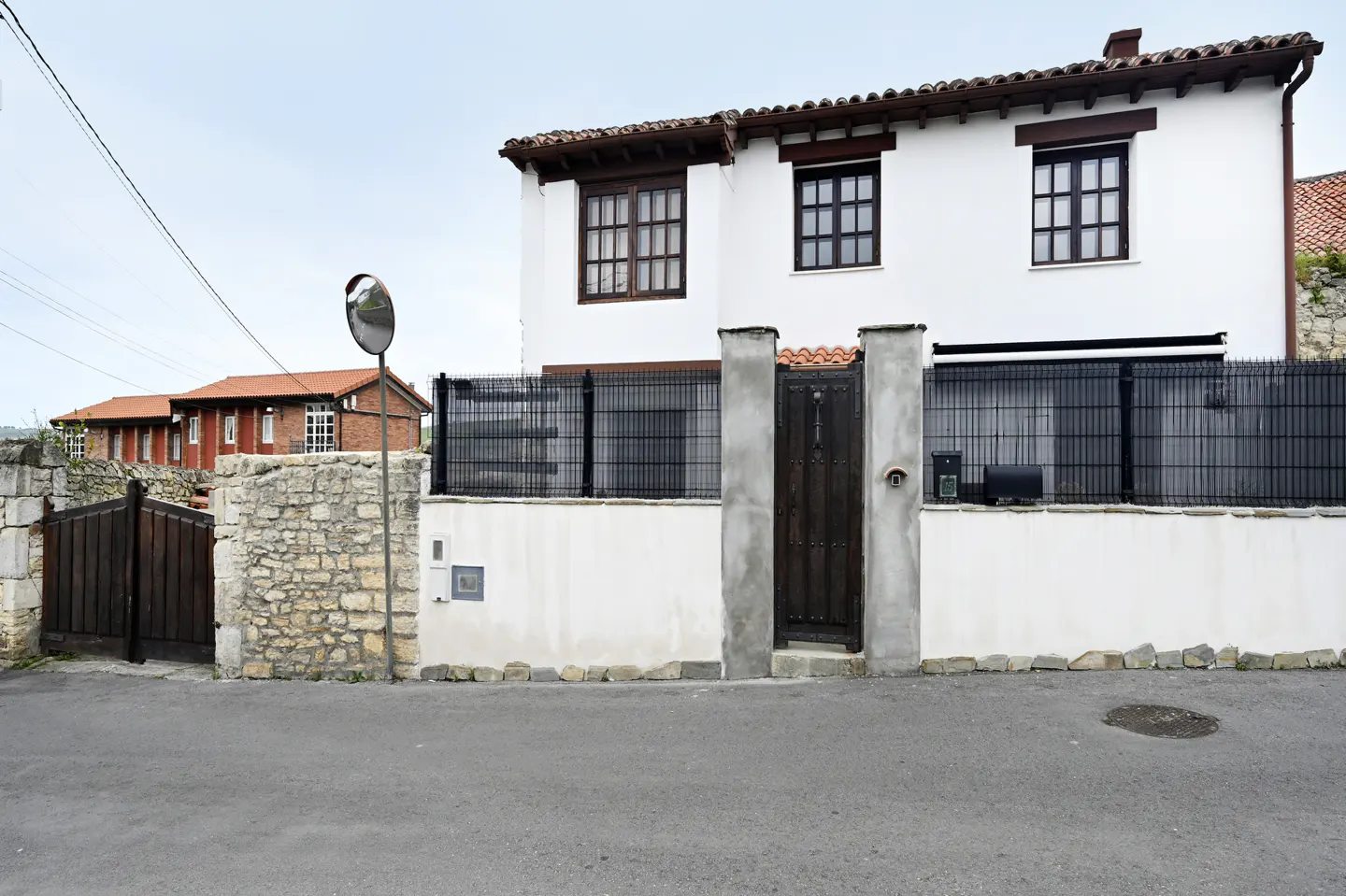Two-story white house with brown trim and a black metal fence. A stone wall and gate are on the left. A traffic mirror is on the side of the road.
