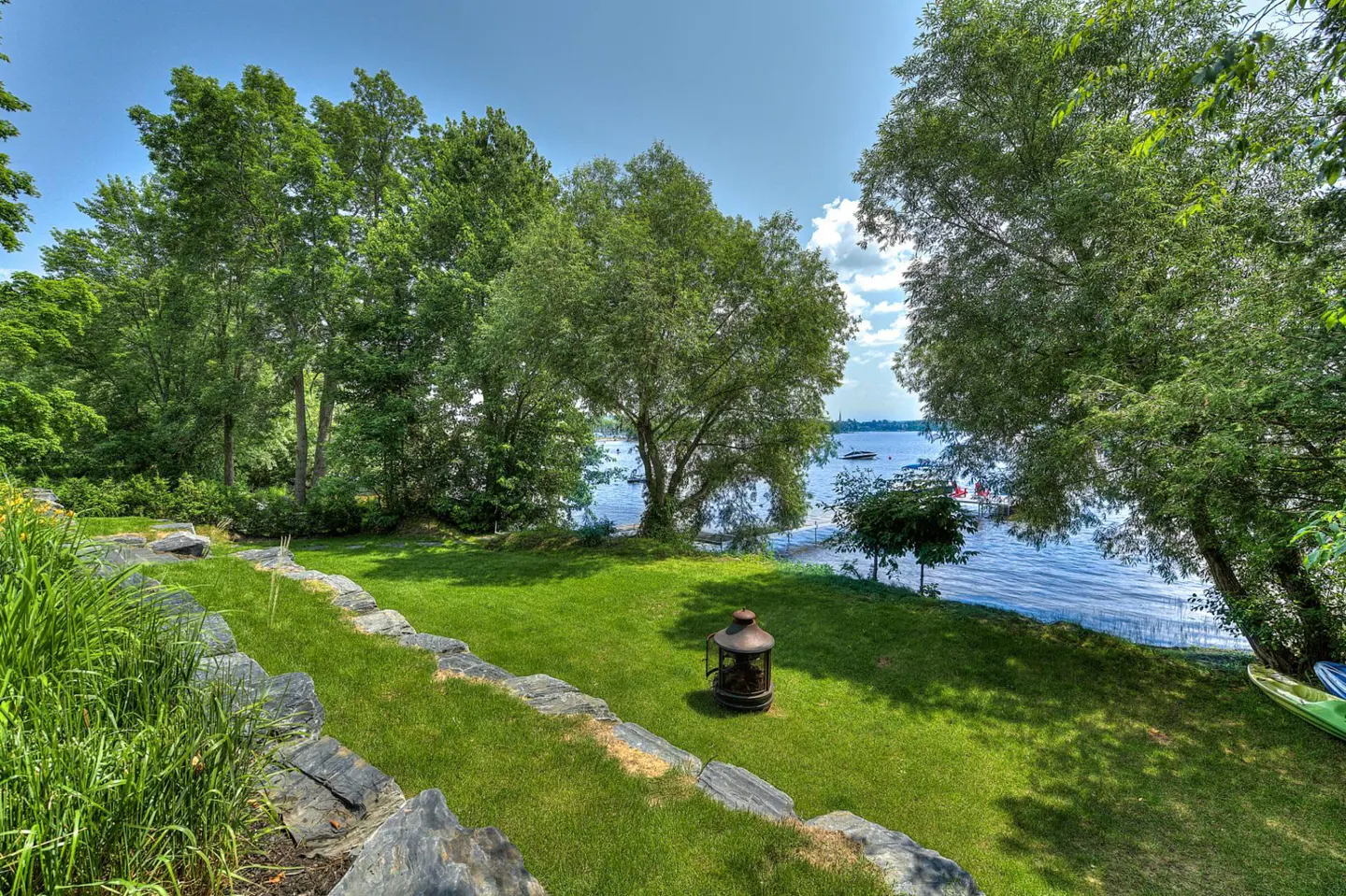 Lawn with stone border slopes to lake. Trees, blue sky, and a metal fire pit are visible.