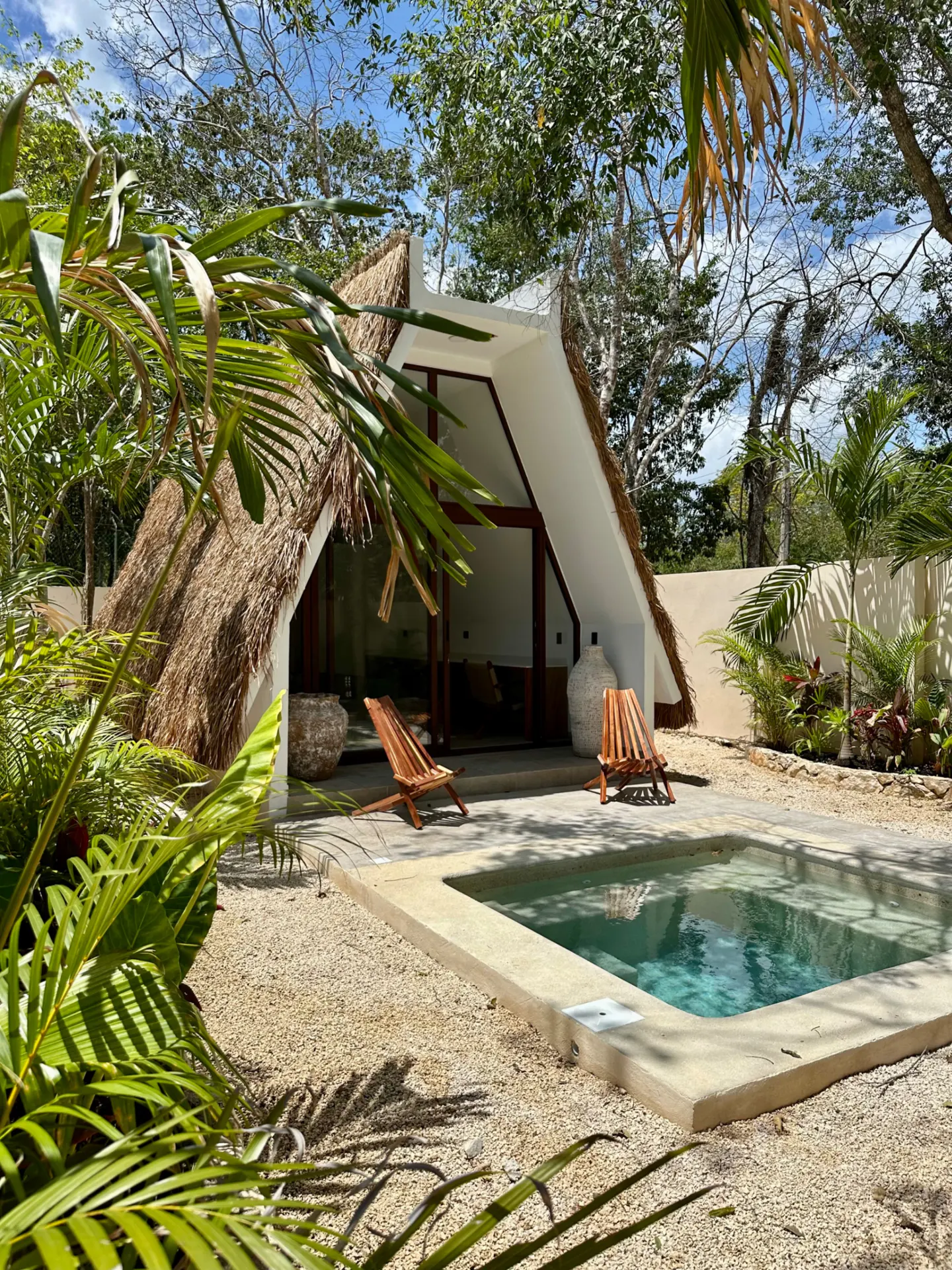 A-frame house with a thatched roof, two wooden chairs, and a small pool surrounded by gravel and lush greenery.