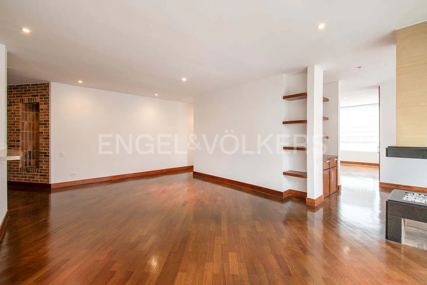 Bright, empty living room with hardwood floors, white walls, and recessed lighting. A brick accent wall and built-in shelves add character.
