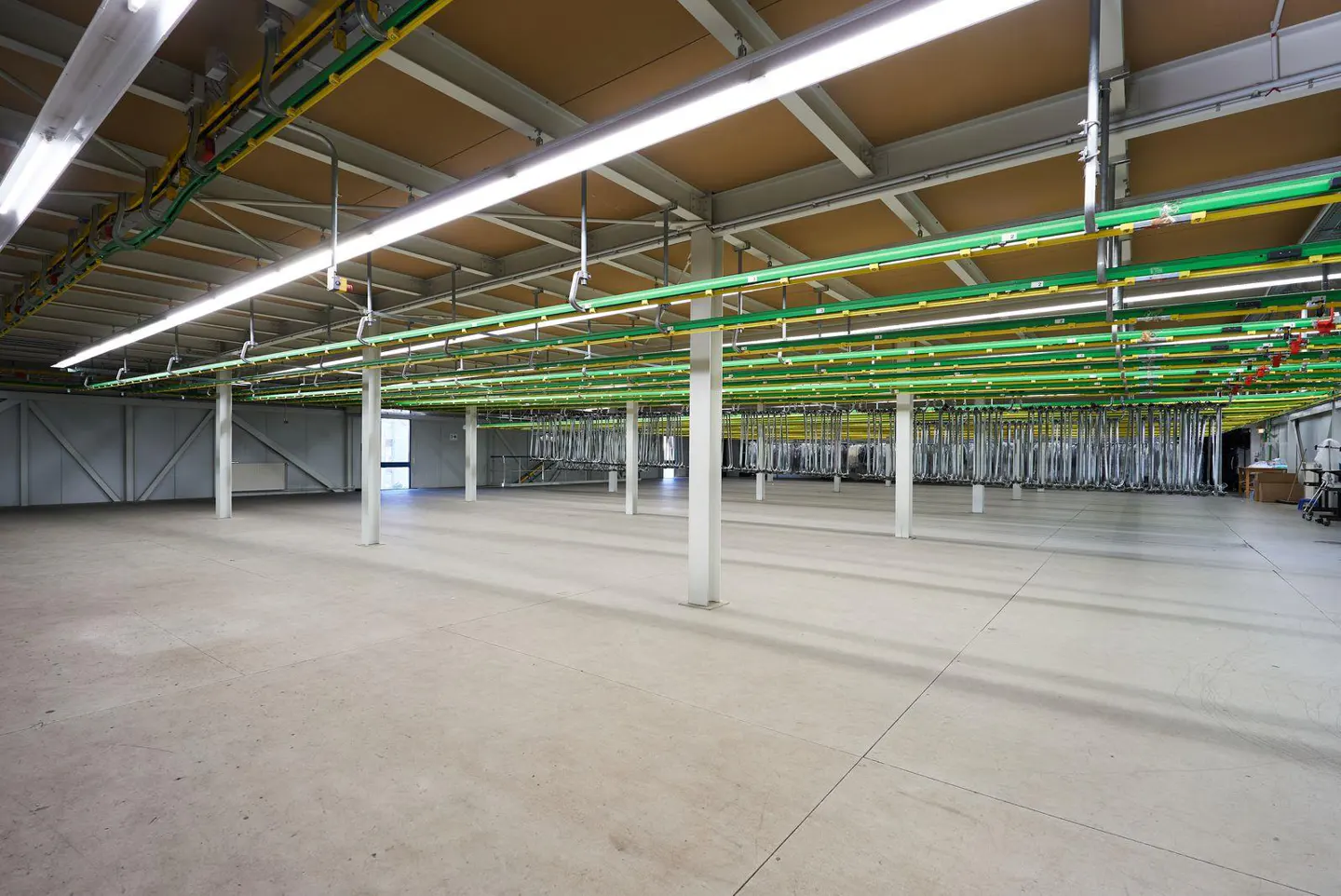 Empty warehouse with concrete floor, white support columns, and green and yellow piping on the ceiling.
