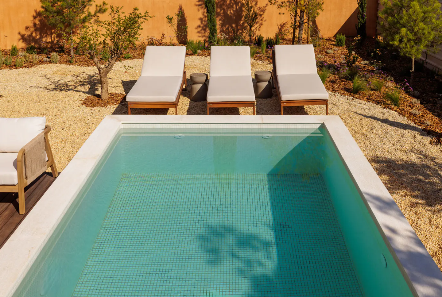 A rectangular pool with turquoise tiles, surrounded by three white lounge chairs and gravel.