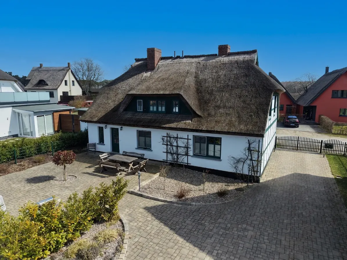 Exterior of a white house with a thatched roof, brick chimney, and a brick-paved courtyard with a wooden picnic table.