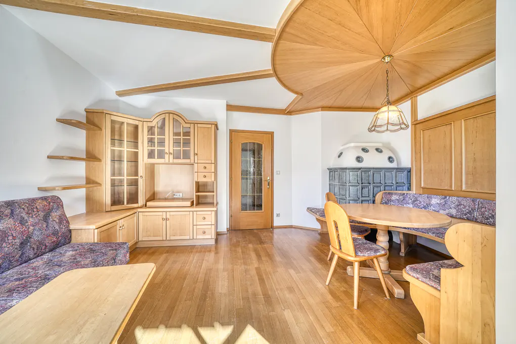 Dining area with wooden furniture, a patterned sofa, a round table, and a traditional tiled stove under a wood ceiling.