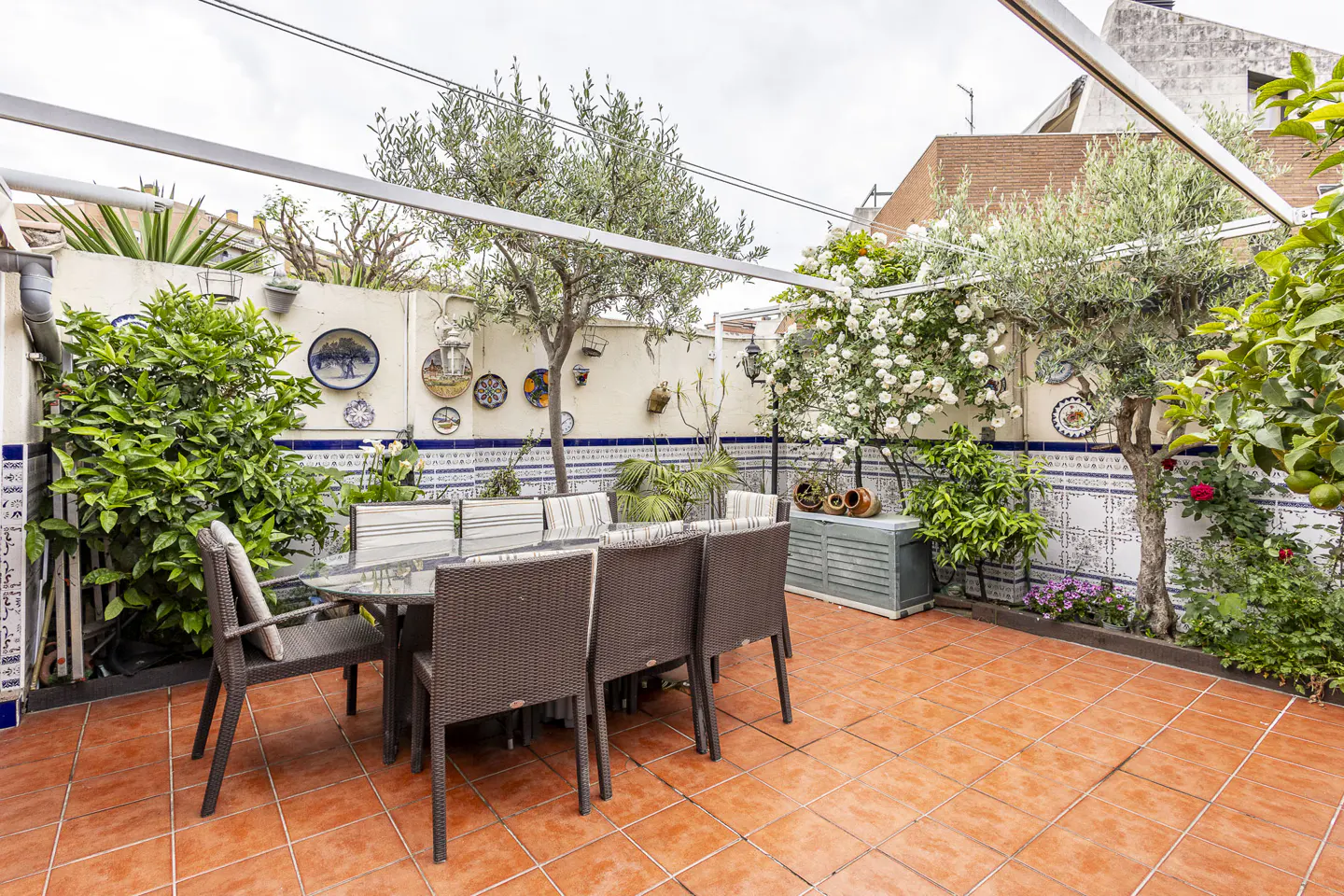 Outdoor patio with a glass table and eight chairs on a terracotta tiled floor, surrounded by plants and a decorated wall.