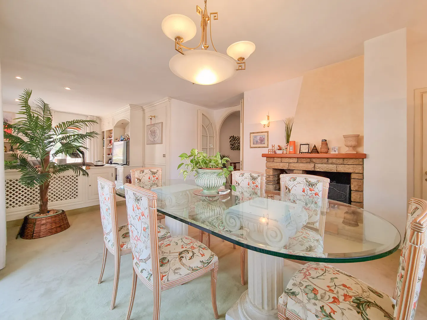Dining room with a glass-topped table, floral chairs, a stone fireplace, and a potted palm tree under a chandelier.
