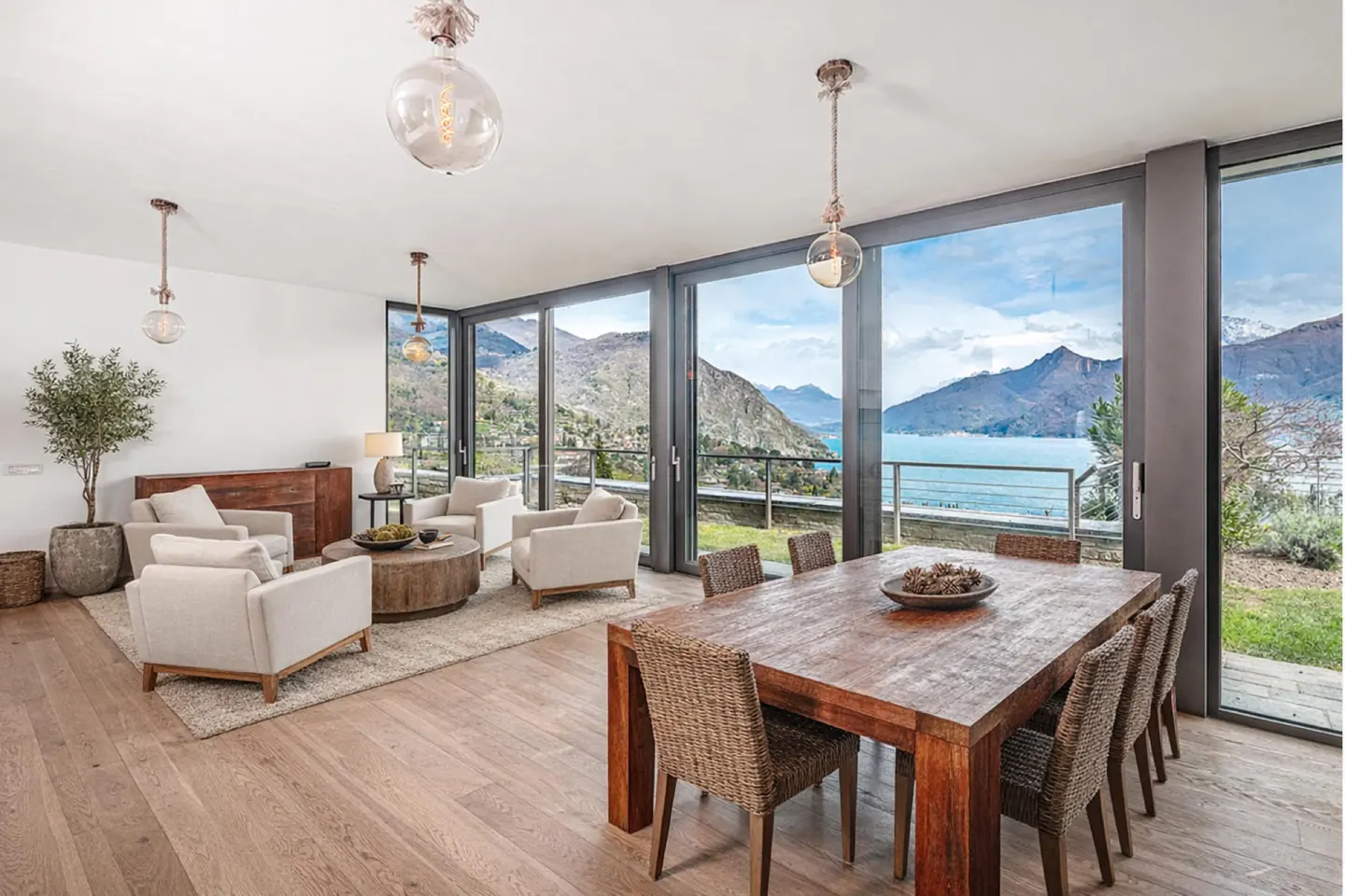 Bright living and dining area with lake view. Wood table with woven chairs, white sofas, and large windows overlooking mountains and water.