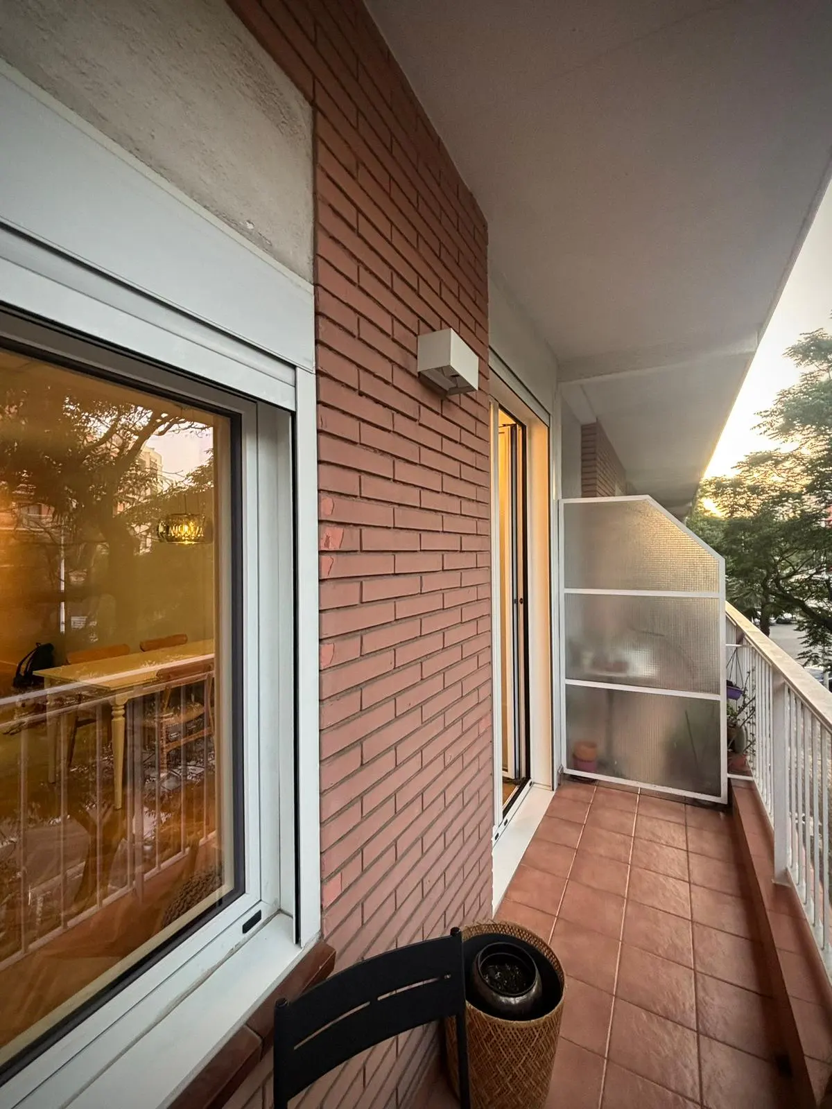 A balcony with red brick walls, terracotta tiles, and a view of trees. A window reflects a dining table.