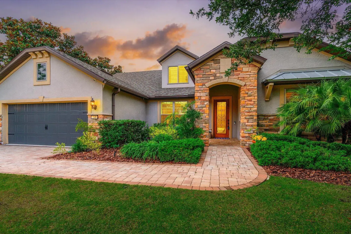 Single-story home with stone accents, a gray garage, and a paved walkway at sunset with lush landscaping.