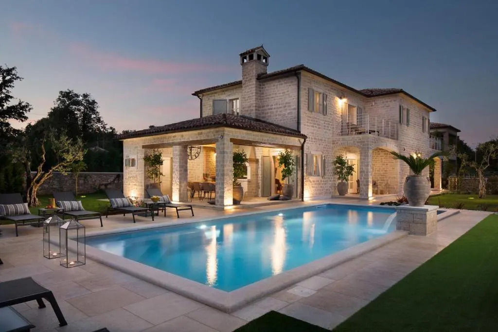 Exterior view of a two-story stone house with a pool at dusk. Lounge chairs and lanterns sit poolside.
