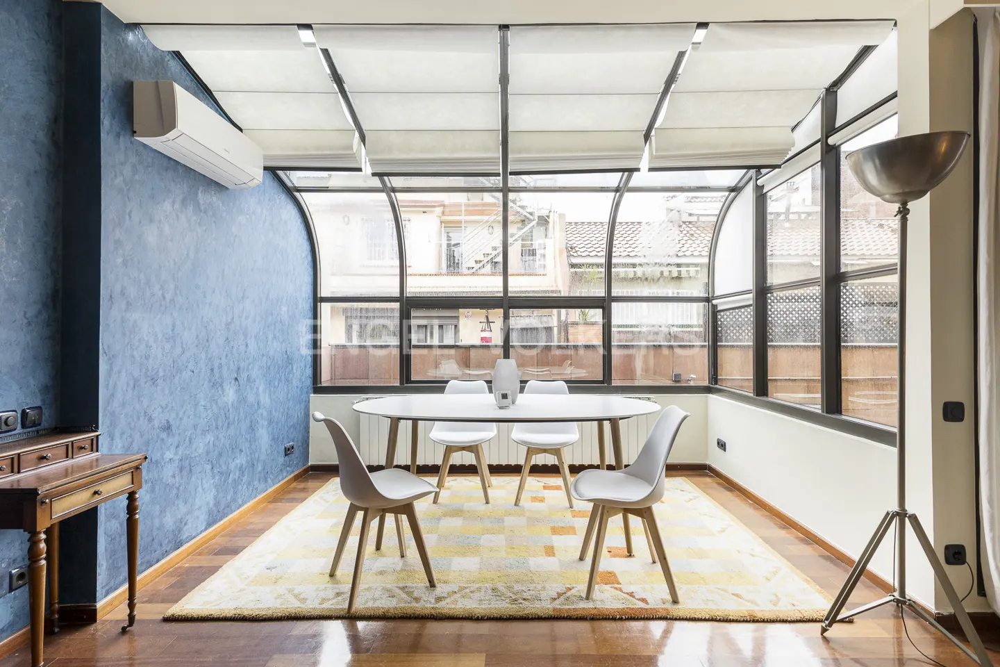 Dining room with blue wall, wood floors, and large windows. An oval table with white chairs sits on a patterned rug. A floor lamp stands near the window.
