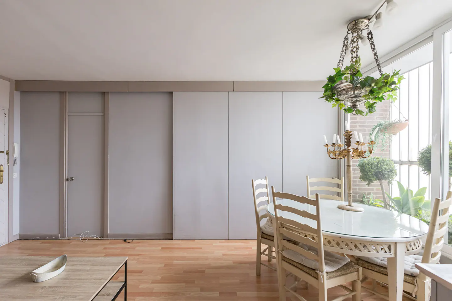 A dining room features a glass-topped table with six chairs, a chandelier with ivy, and a large window with plants.
